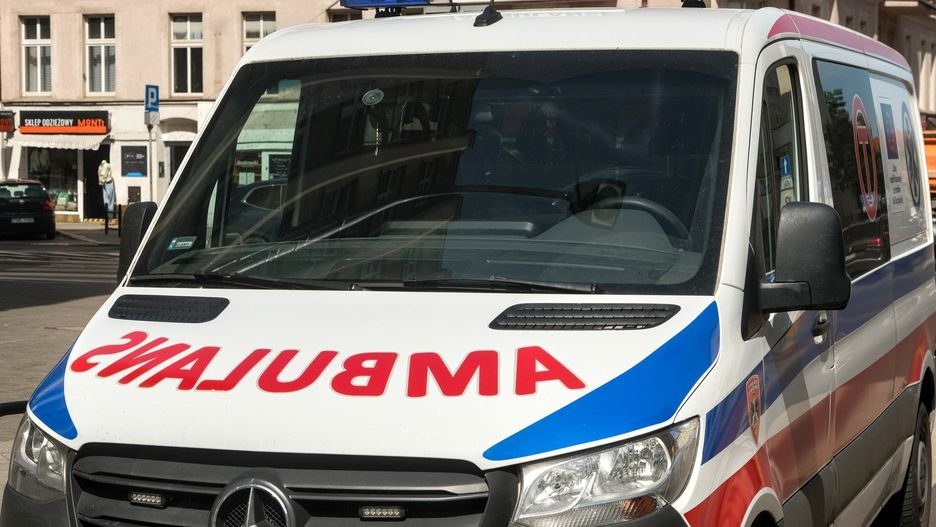 White ambulance van with red â€œAMBULANSâ€ sign and blue emergency lights , parked on a city street near shops and buildings.