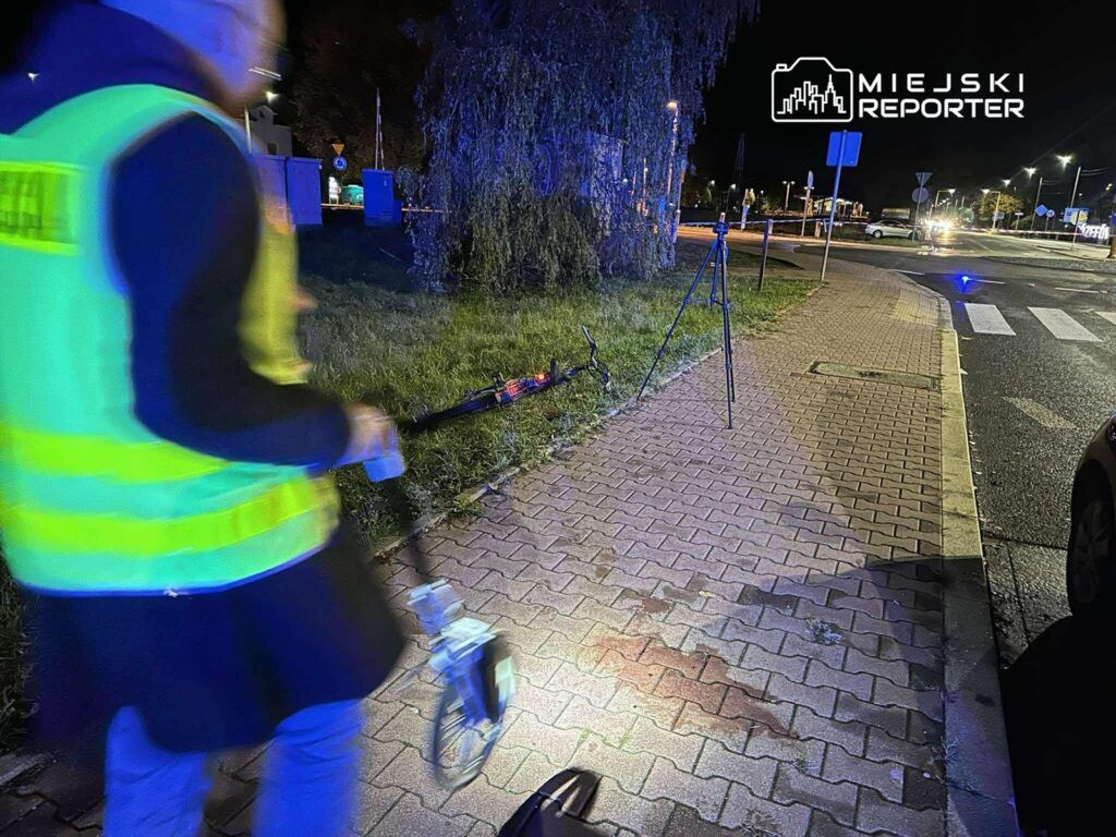 Mężczyzna w odblaskowej kamizelce mierzy odległość przy ulicy nocą, obok statywu i nieoświetlonego terenu.