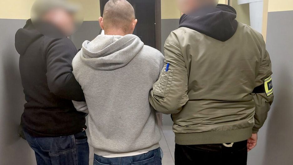 Mężczyzna w szarej bluzie jest prowadzony przez dwóch funkcjonariuszy w mundurach w korytarzu budynku.