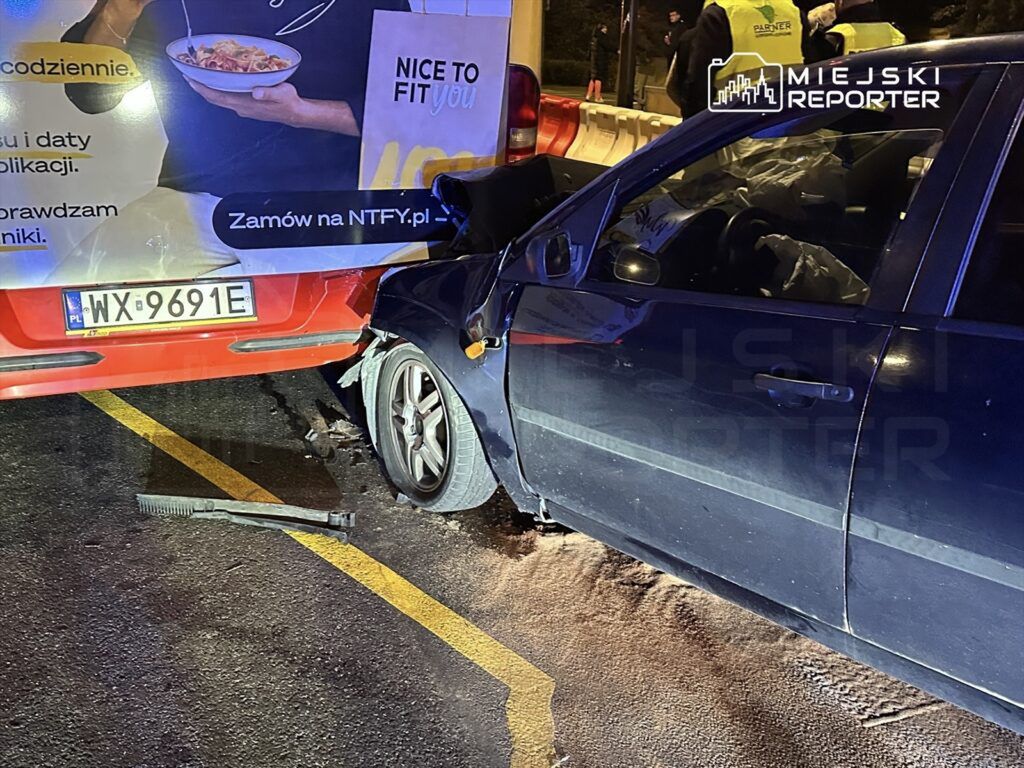 Czarny samochód osobowy zderzony z czerwoną furgonetką na ulicy, obok leżący przedmiot przypominający narzędzie.