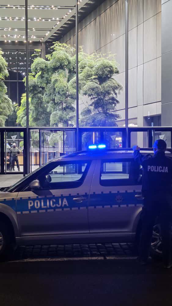 Policjant przy radiowozie z włączonymi sygnałami świetlnymi, stojący przed nowoczesnym budynkiem.
