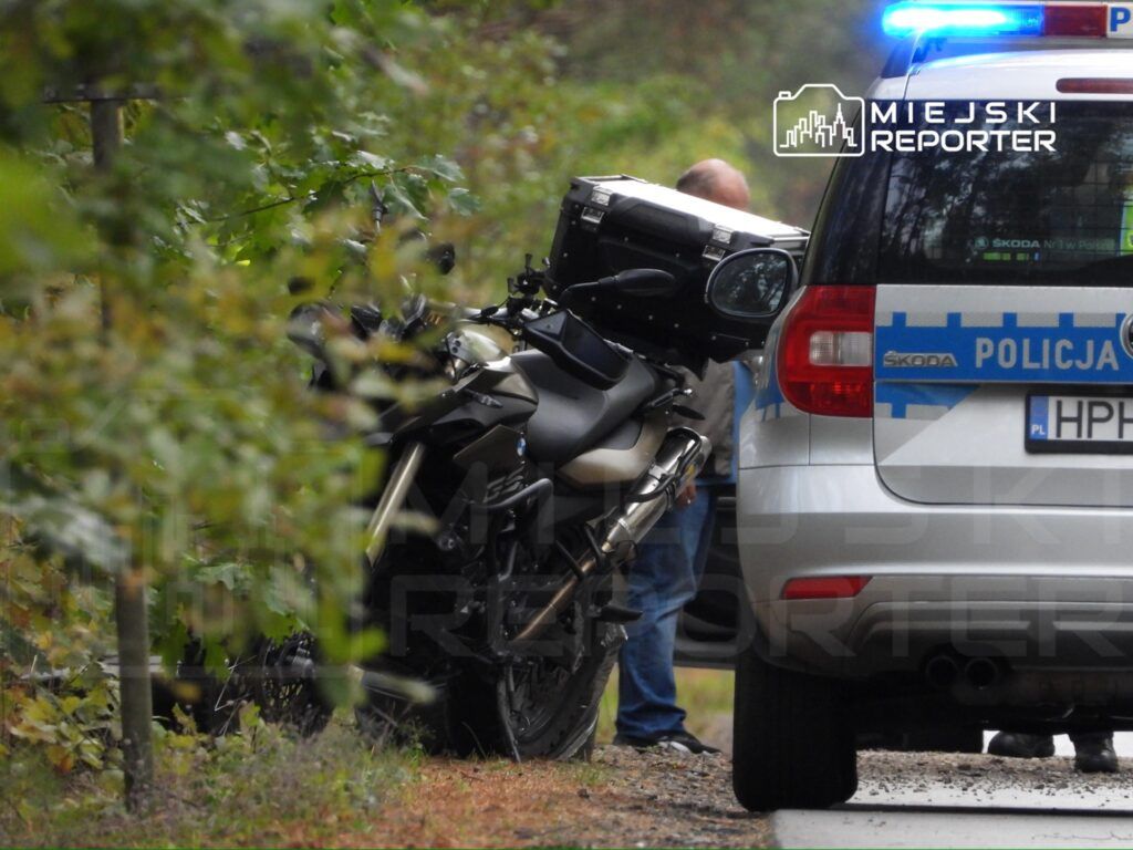 Motocyklista w zbroi stoi obok policyjnego samochodu z włączonymi sygnałami świetlnymi na leśnej drodze.