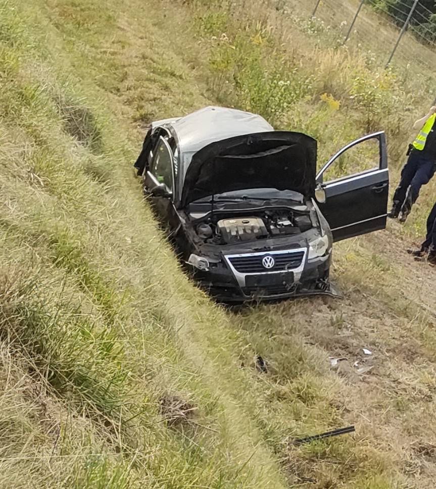 Czarny samochód osobowy marki Volkswagen stoi w rowie, z otwartą maską, obok niego stoi funkcjonariusz w kamizelce odblaskowej.