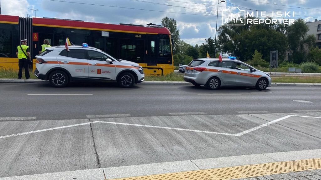 Funkcjonariusze w kamizelkach odblaskowych kontrolują ruch obok tramwaju na ulicy, obok zaparkowane pojazdy ZTM.