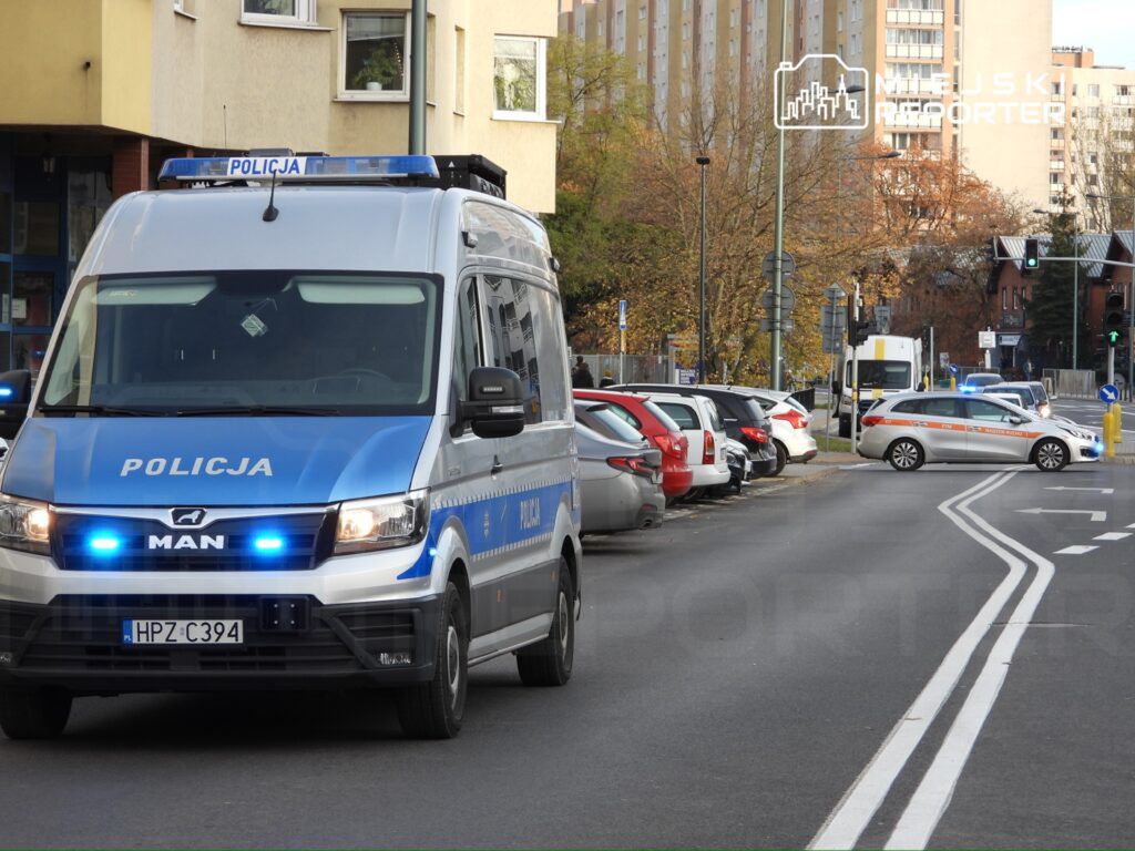 Radiowóz policyjny z włączonymi sygnałami świetlnymi zatrzymany na ulicy w pobliżu zaparkowanych samochodów.