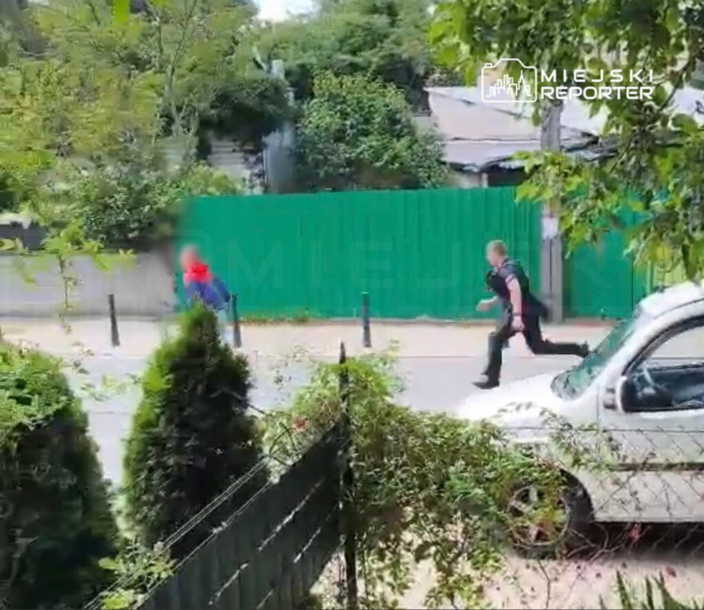 Mężczyzna w niebieskiej kurtce i czerwonej czapce biegnie ulicą, a za nim podąża policjant w mundurze.