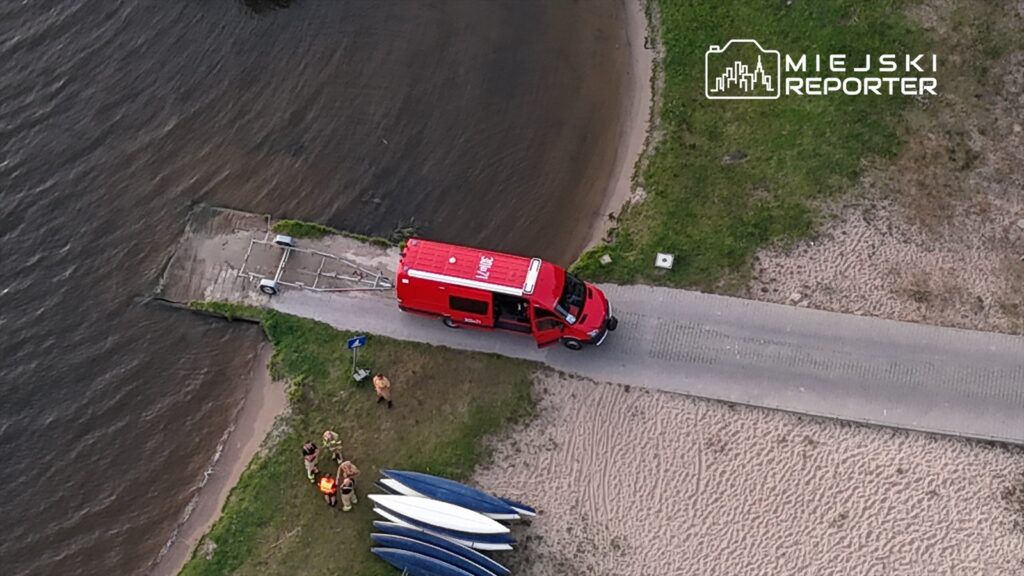 Czerwony pojazd ratunkowy stoi na drodze obok wody, a grupa ratowników przygotowuje sprzęt na plaży.