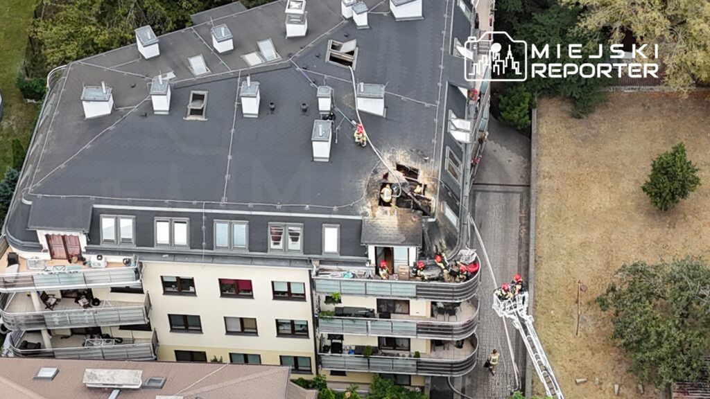 Grupa strażaków pracuje nad usuwaniem skutków pożaru na dachu nowoczesnego budynku mieszkalnego.