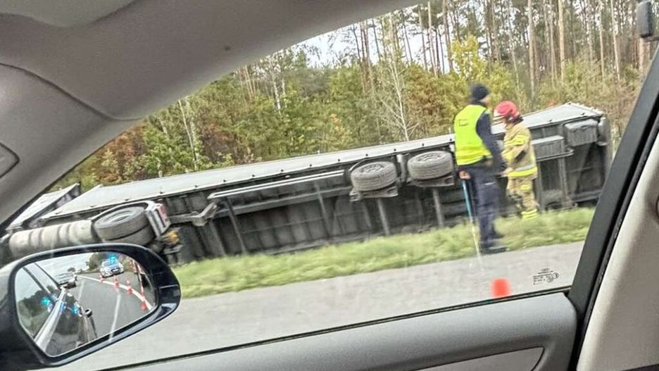 Strażak i policjant rozmawiają obok przewróconego ciężarowego pojazdu na poboczu drogi, w otoczeniu lasu.
