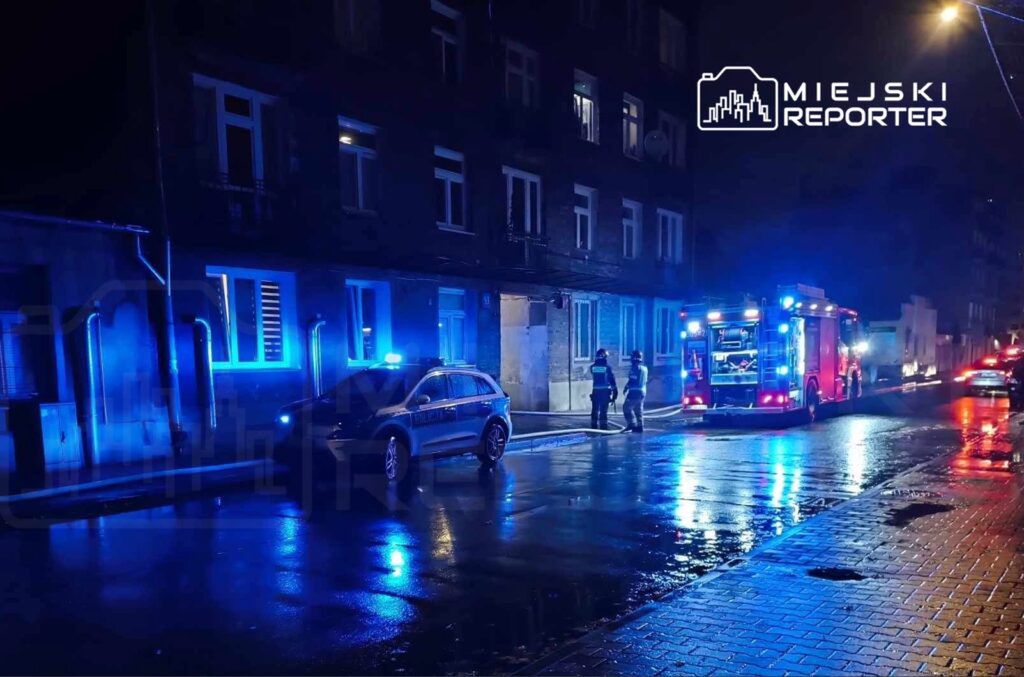 W nocy, w deszczowej scenerii, strażacy w mundurach rozmawiają obok wozu strażackiego i radiowozu policyjnego.