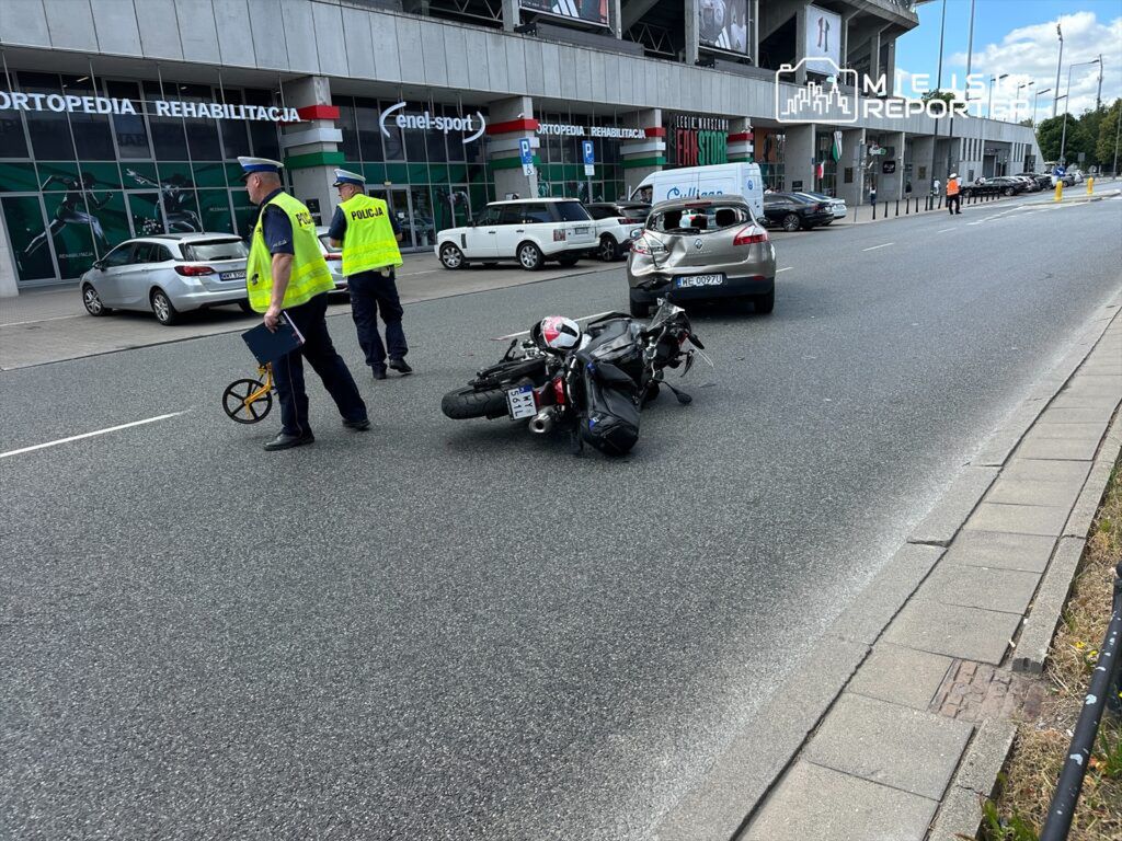 Policjanci prowadzący czynności dochodzeniowe przy przewróconym motocyklu na ulicy obok nowoczesnego biurowca.