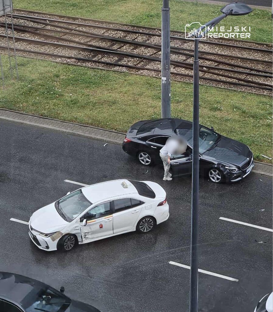 Czarny samochód osobowy stoi na jezdni, a obok niego mężczyzna sprawdzający uszkodzenia białego taxi po kolizji.