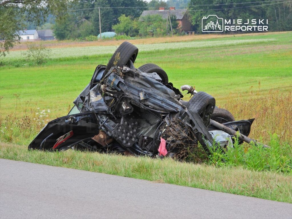 Przewrócony samochód leży na boku w zaroślach obok drogi, otoczony zielonymi polami i drzewami.