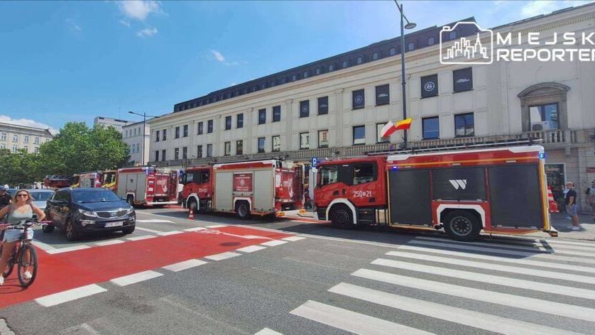 Grupa strażaków w wozach strażackich blokuje ulicę w centrum miasta, podczas gdy przechodnie i rowerzyści czekają na możliwość przejścia.