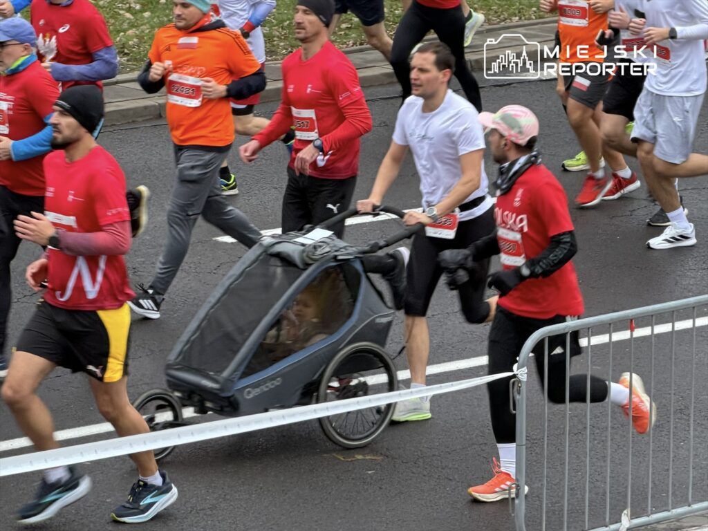Biegacze w kolorowych strojach uczestniczą w biegu ulicznym, a jeden z nich pcha wózek dziecięcy.