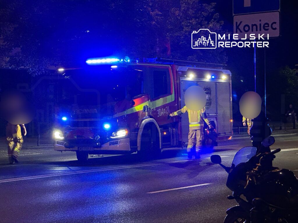 Strażacy w mundurach zbliżają się do wozu strażackiego na oświetlonej ulicy nocą.