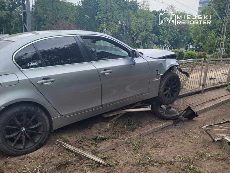 Uszkodzony srebrny samochód osobowy stoi na krawędzi chodnika, z jednym kołem na drewnianej belce, otoczony zielenią.
