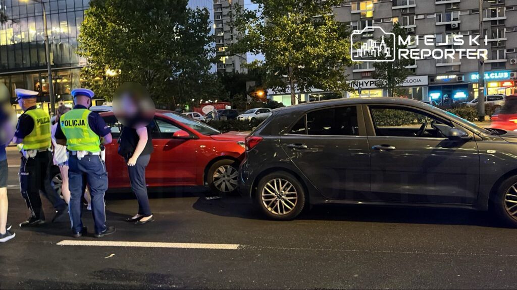 Policjanci rozmawiają z kobietą przy zderzonych samochodach na ulicy w mieście, w tle widoczne oświetlone budynki.