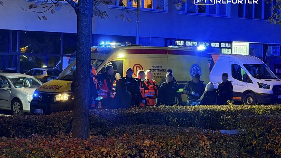 Grupa ratowników medycznych z ambulansu interweniuje w nocy przy budynku mieszkalnym, otoczona przez krzewy.