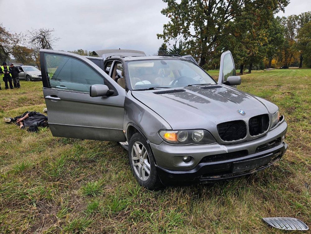 Uszkodzony samochód osobowy marki BMW stoi na trawie, z otwartymi drzwiami. W tle widać funkcjonariuszy policji.