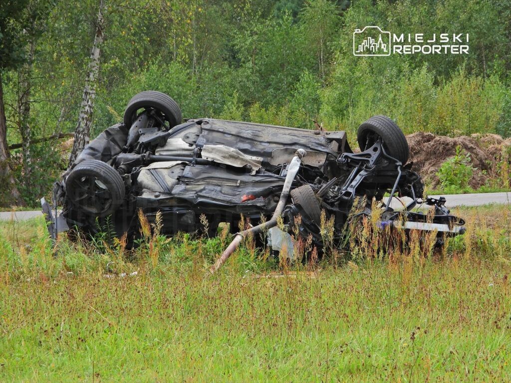 Przewrócone samochód osobowy leży na boku w wysokiej trawie przy drodze, otoczone drzewami.