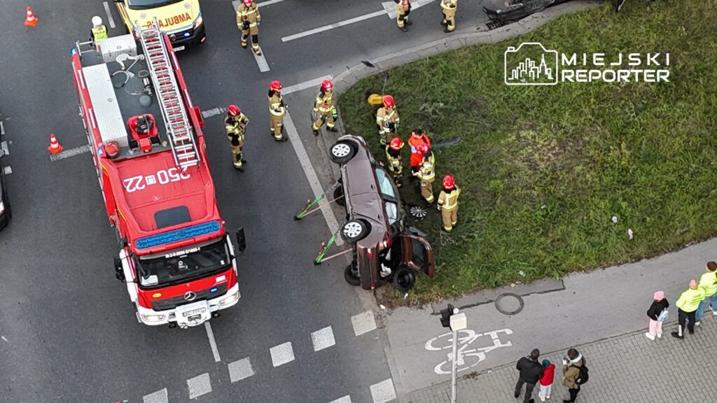 Wielu strażaków pracuje przy przewróconym samochodzie na poboczu drogi, obok straży pożarnej i ambulansu.