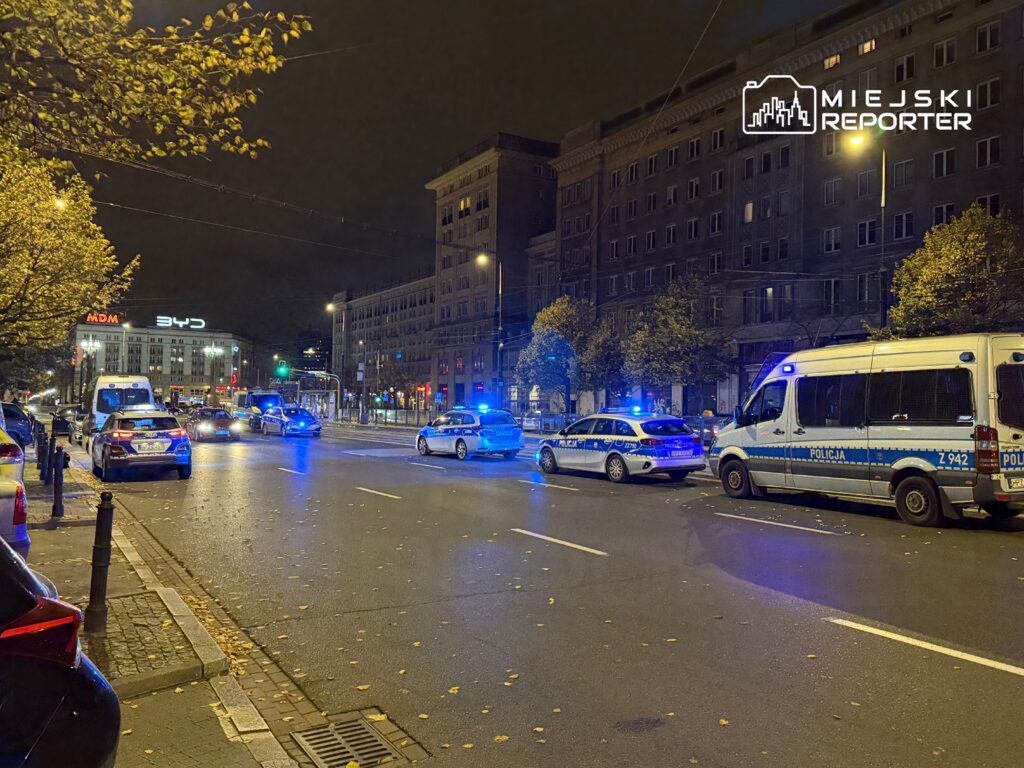 Policyjne samochody z włączonymi sygnałami świetlnymi zatrzymane na ruchliwej ulicy nocą, w tle oświetlony budynek.