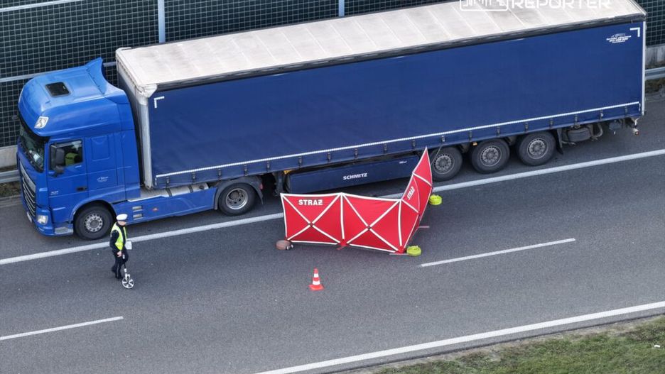 Policjant w kamizelce odblaskowej mierzy odległość obok niebieskiego tira, a w tle znajduje się czerwony namiot strażacki.