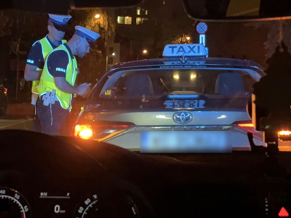 Dwaj policjanci w kamizelkach odblaskowych kontrolują taksówkę na ulicy w nocy.