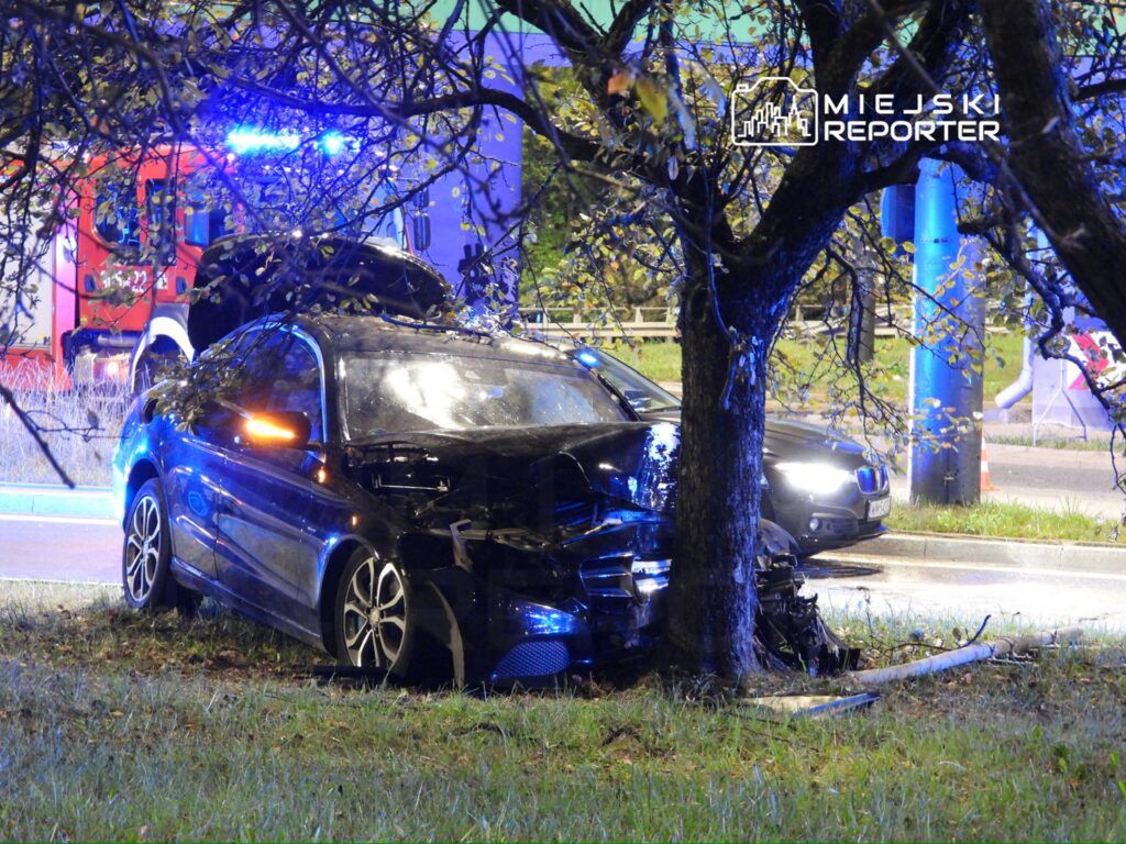 Czarny samochód osobowy uderzony w drzewo, obok widoczny wóz strażacki z włączonymi sygnałami świetlnymi.