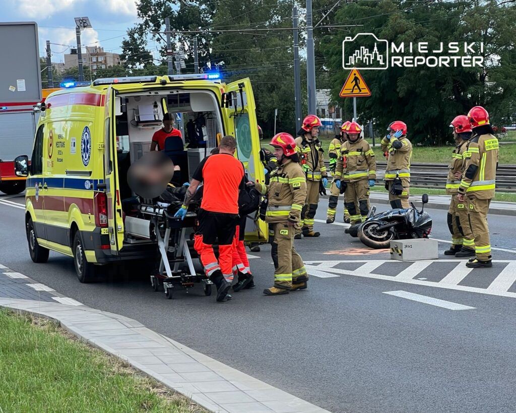 Ratownicy medyczni transportują poszkodowanego na noszach do karetki, obok leży przewrócony motocykl.