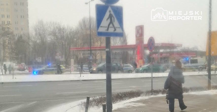 Mężczyzna w szarej kurtce stoi na chodniku obok znaku drogowego, w tle widać pojazdy policyjne i stację benzynową.