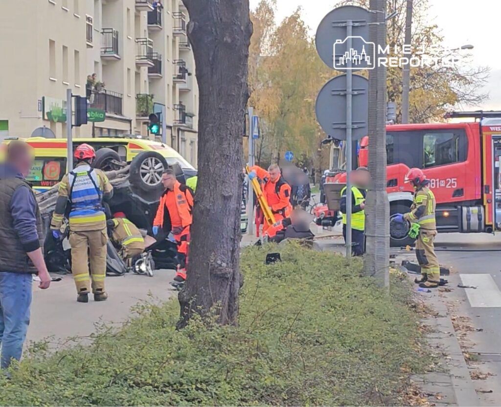 Grupa ratowników medycznych i strażaków udziela pomocy przy przewróconym samochodzie na ulicy, obok sygnalizacji świetlnej.