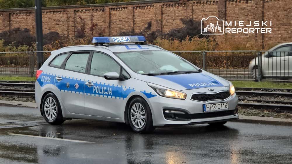 Policyjny samochód osobowy zaparkowany na ulicy obok torów tramwajowych w deszczowy dzień.