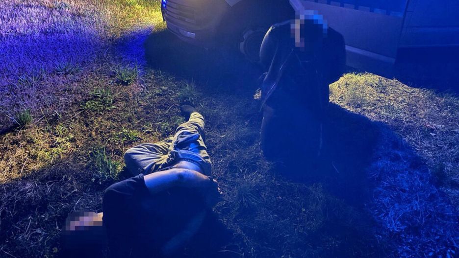 Dwóch mężczyzn leży na trawie obok radiowozu policyjnego, oświetlonych niebieskimi światłami.