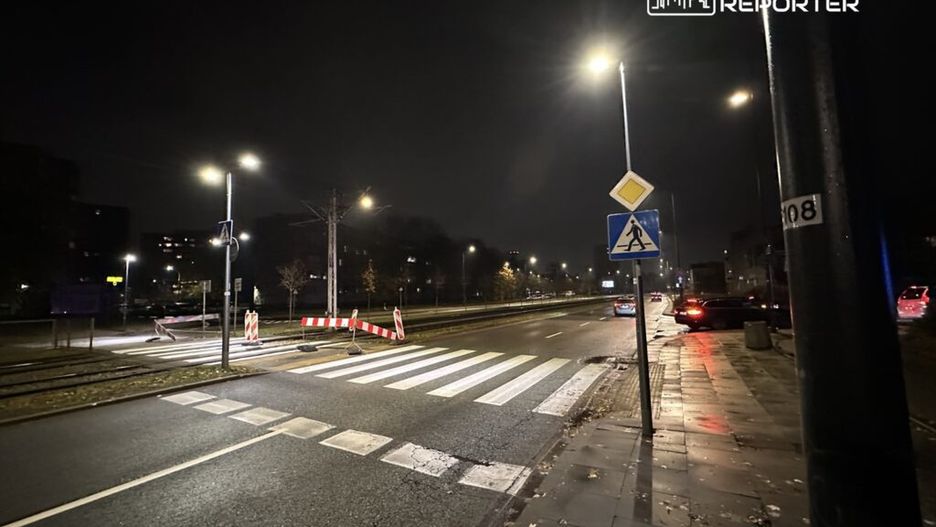 Oświetlona ulica nocą z przejściem dla pieszych, barierkami drogowymi i zaparkowanymi samochodami.