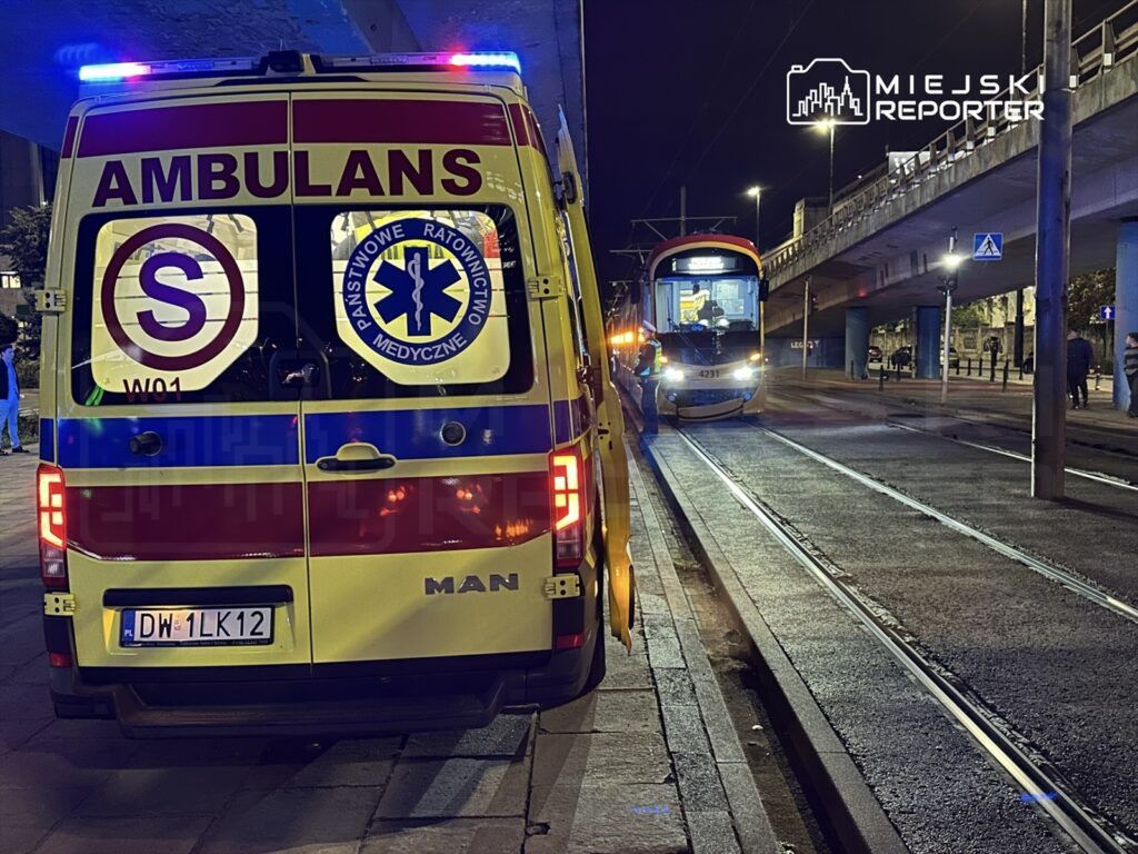 Ambulans z włączonymi sygnalizacjami świetlnymi stoi na ulicy obok tramwaju w nocy.