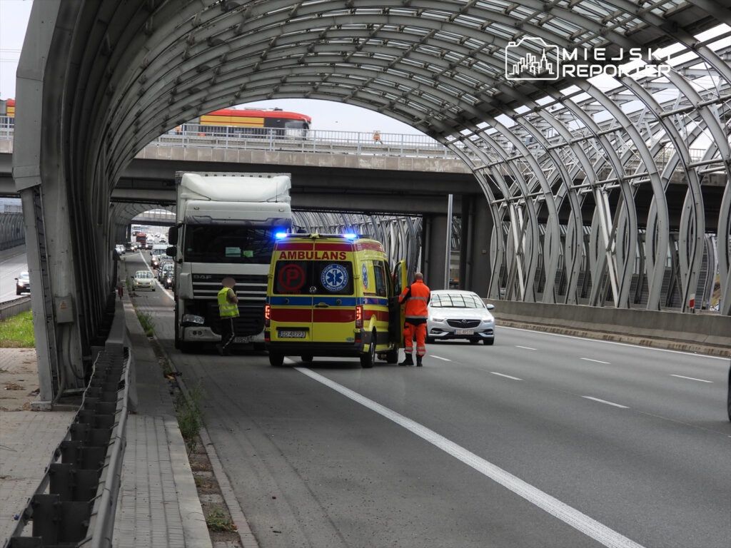 Ambulans z włączonymi sygnałami zatrzymany obok ciężarówki na drodze, obok pracownik w kamizelce odblaskowej.
