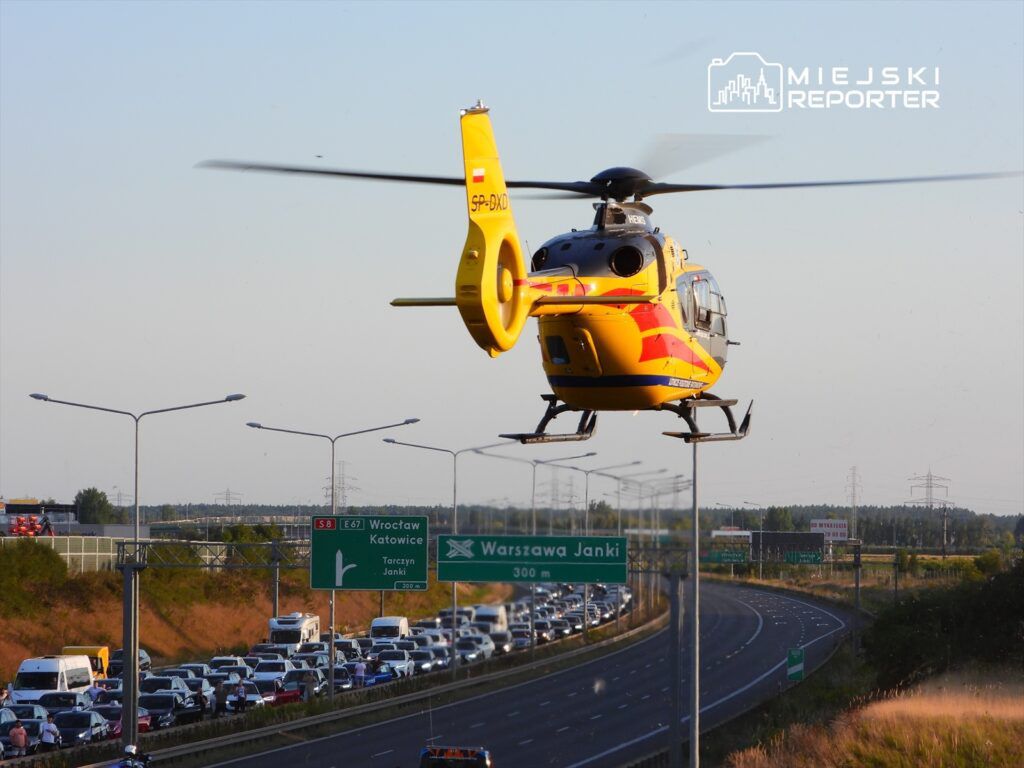 Żółty helikopter przelatuje nad zakorkowaną autostradą, z widocznymi znakami drogowymi wskazującymi kierunek do Warszawy i Katowic.