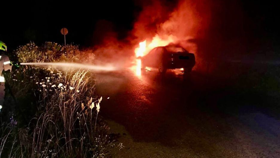 Strażak polewa wodą płonący samochód na drodze w nocy, otoczony dymem i wysoką trawą.