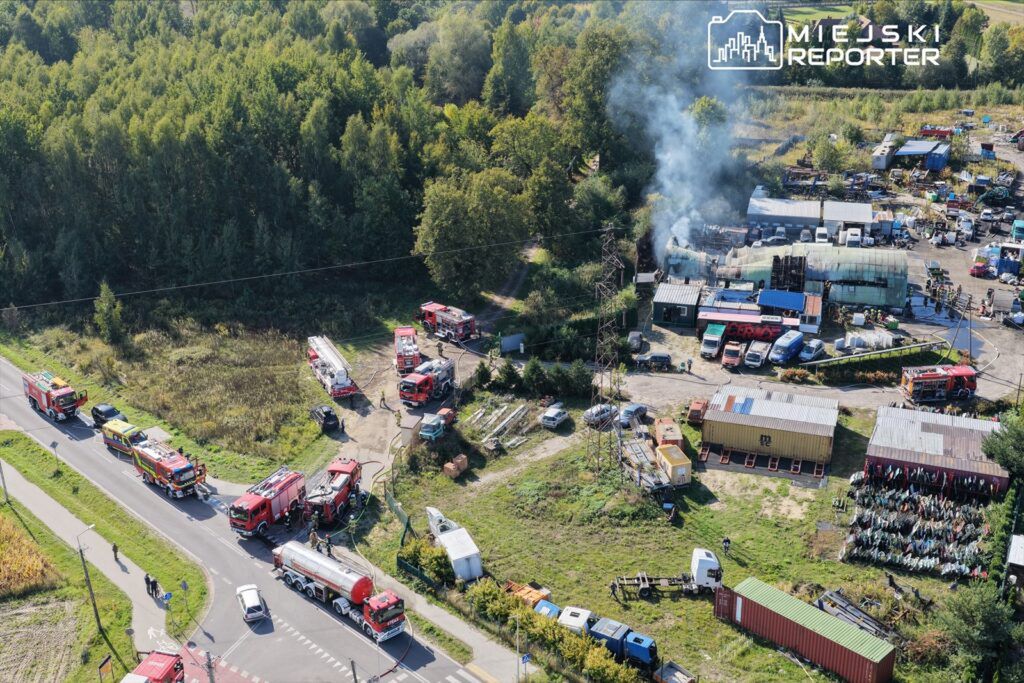 W okolicy zakładu przemysłowego strażacy pracują nad gaszeniem pożaru, a w tle unosi się dym.
