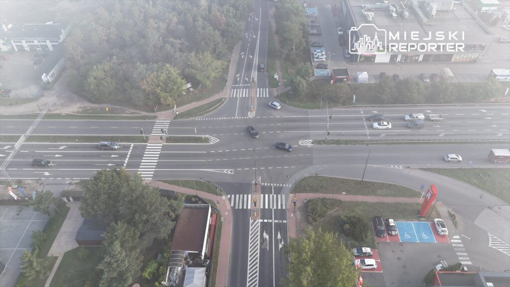 Ruchliwa skrzyżowanie z samochodami poruszającymi się w różnych kierunkach, otoczone drzewami i parkingiem.