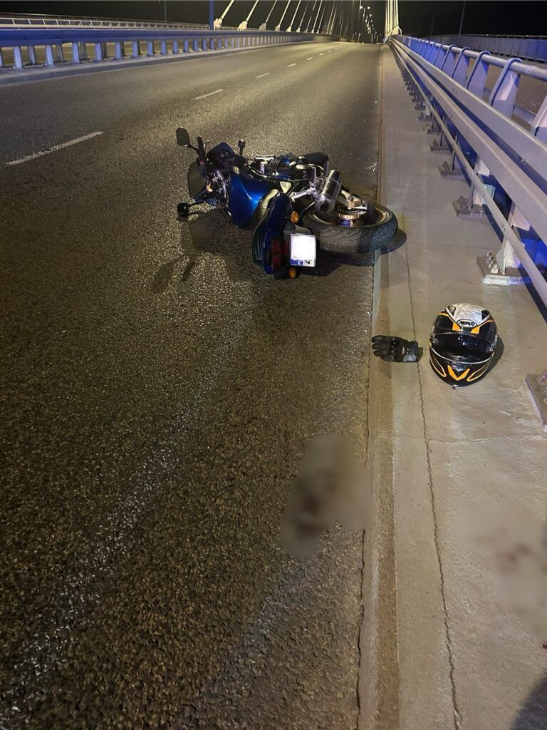 Motocykl leży na jezdni na moście, obok znajduje się kask i rękawica. Nocna sceneria z oświetleniem.