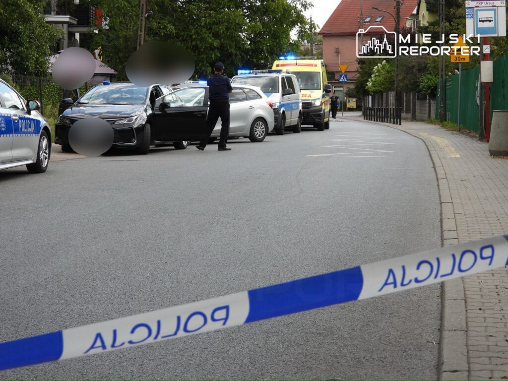 Policyjny radiowóz i karetka stoją na ulicy, a policjant rozmawia z kierowcą czarnego samochodu.