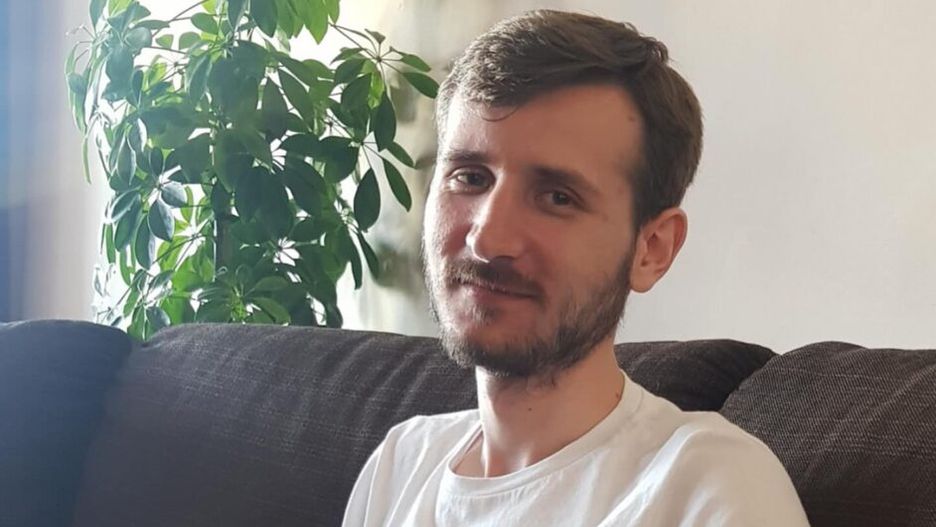 Mężczyzna w białej koszulce siedzi na kanapie z psem, trzymając go na kolanach, w tle rośliny doniczkowe.