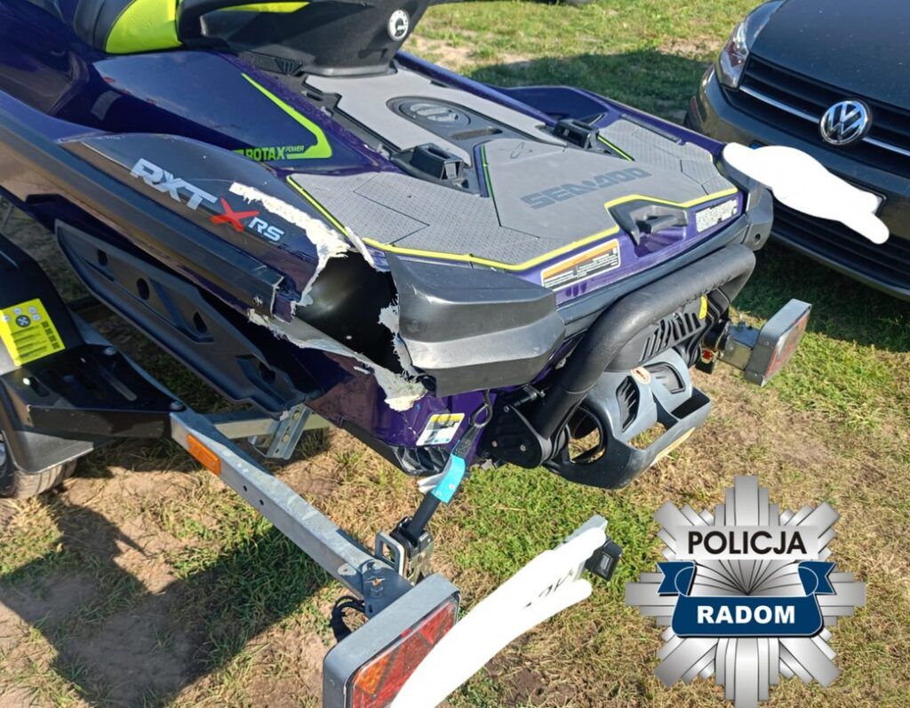 Uszkodzona motorówka wodna zaparkowana na przyczepie w otoczeniu trawy i samochodu osobowego.
