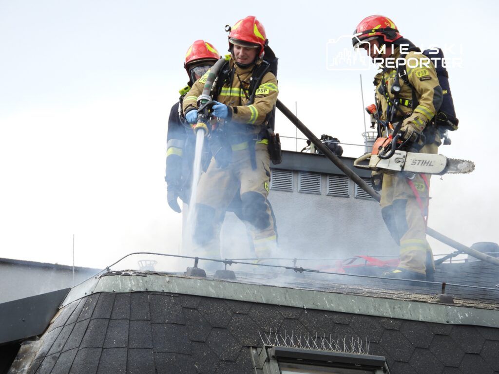 Strażacy gaszą ogień na dachu budynku, używając węża gaśniczego oraz piły łańcuchowej, w otoczeniu dymu.