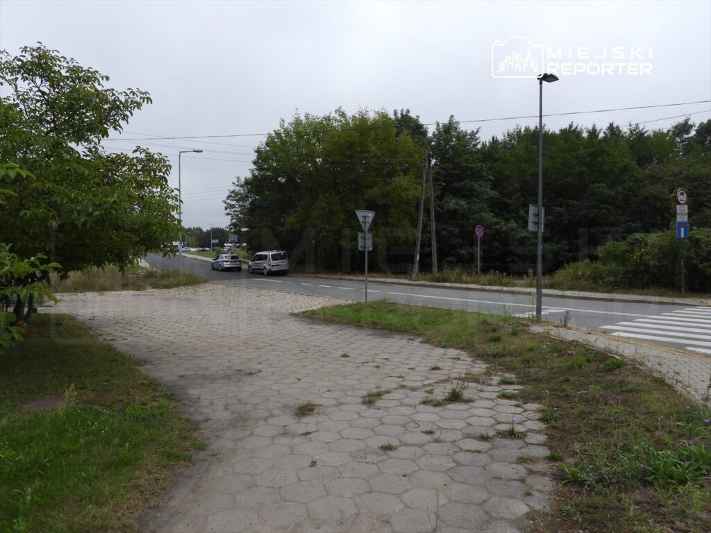 Zielony skwer z kostką brukową prowadzi do ruchliwej drogi, gdzie widoczne są dwa samochody. W tle drzewa i znaki drogowe.