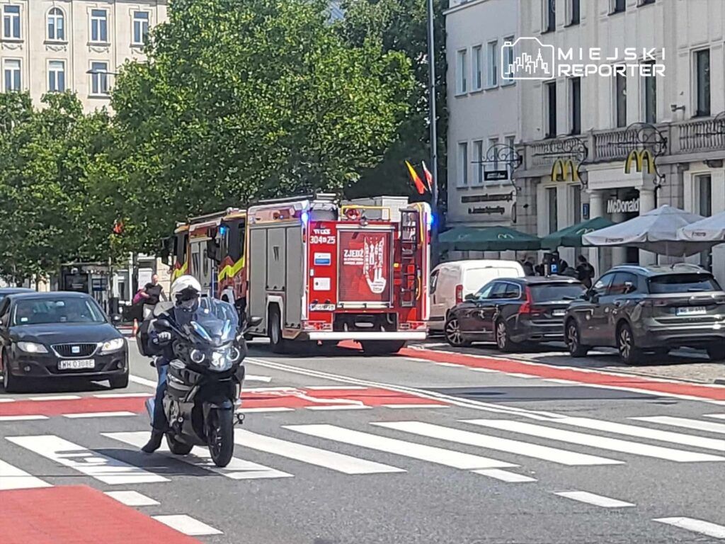 Motocyklista przejeżdża obok wozu strażackiego z włączonymi sygnałami świetlnymi na ulicy z zielonymi drzewami w tle.