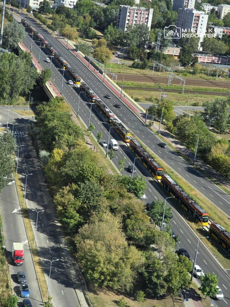 Długi szereg tramwajów kursujących po torach obok ruchliwej ulicy w otoczeniu zieleni i zabudowy miejskiej.